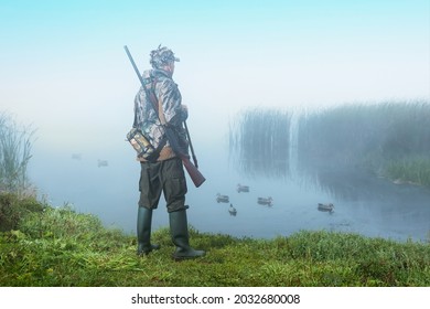 Hunter With Gun Stands On The Shore Of Lake At Sunrise. Hunting With Ducks Decoy.