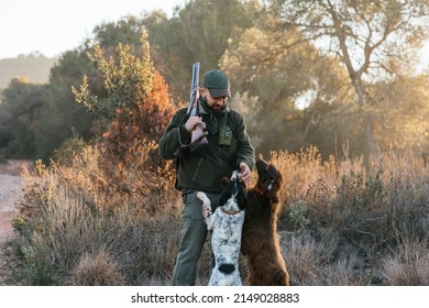 Hunter with his shotgun on shoulder petting his two dogs in nature