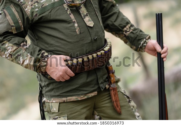 Hunter man stands with a belt of ammunition and a gun in\
his hands. A hunter with cartridges on his belt stands in the\
forest. 