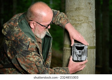 Hunter sets a trail camera on a tree in the forest. Trail cameras are often used by hunters for automatic photography or video shooting of wildlife in the forest. - Shutterstock ID 2324851003