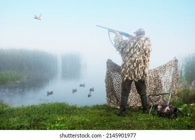 Hunter Shooting Into Sky During Duck Hunting At Sunrise. Hunting With Ducks Decoy On River Bank.