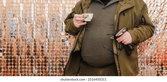 Hunter's outfit for the fall season; An elderly man drinks coffee while taking a break from hunting, holding glasses and gloves in his other hand; the background sparkles. - Powered by Shutterstock