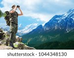 hunting, army, military service and people concept - young soldier with backpack looking to binocular over mountains observing area