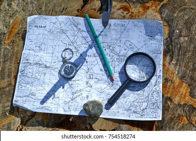 Hunting Folding Knife With Steel Sharp Blade Stuck On Old Tree Stump In The Wild Forest Pressing Compass, Magnifying Glass, Vintage Military Topographic Map Of The Area, Pencil, Stone