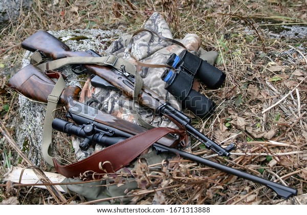 Hunting rifles and binoculars lie on a camouflage\
jacket. Old Soviet and new German hunting weapons with an optical\
sight on the grass. 