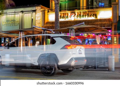 Huntington Beach, CA / USA - February 29, 2020: People Having A Fantastic Time At Restaurants And Bars On Saturday Night On Main Street, With Long Exposure Creating Light Trails And Transparent Car.