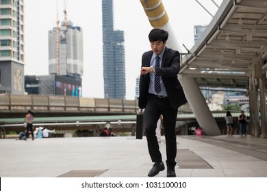 Hurry Asian Businessman Running And Looking At Watch To Check Time On City Walk In Rush Hour. Young Man Late For Train Transportation, Work, Meeting. Office Life And Business Competition.