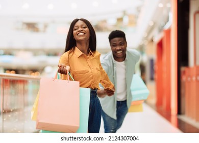 Hurry Up. Portrait Of Excited African American Woman Pulling Guy To Buy New Clothes And Gifts, Fooling And Running In Shopping Mall Together Holding Bags. Discounts, Sales Purchase And Retail Concept