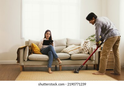 Husband calmly does housework vacuuming the carpet in the living room. while his wife sits comfortably on the sofa playing on her laptop interested in social networks don't want to do housework. - Shutterstock ID 2190771239