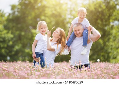 A husband with his wife and two children play in a field of pink flowers. Man, woman and children are wearing white T-shirts and jeans.