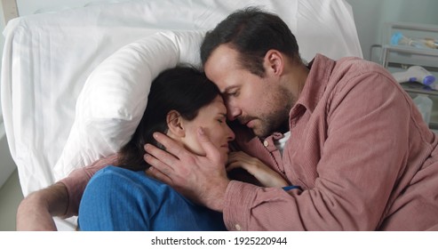 Husband Hugging And Comforting Crying Sick Woman Lying Together In Hospital Bed. Man Visiting Ill Wife Patient Feeling Scared And Depressed Resting In Ward