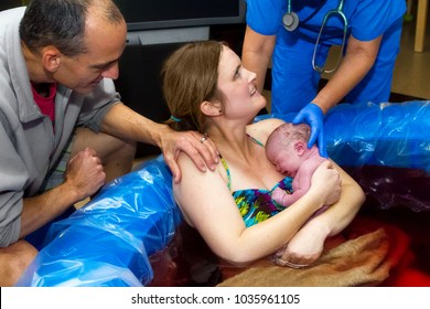 A husband and a midwife both congratulate a new mother.  Their supportive hands are on her shoulder and the back of the newborn baby head, moments after birth.