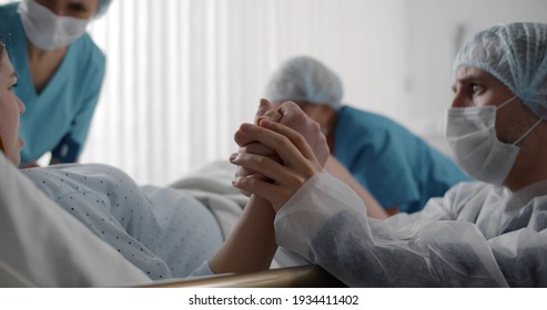 Husband In Protective Mask Comforting Pregnant Woman During Labor In Ward Of Hospital. Young Woman Pushing Giving Birth In Clinic Delivery Room With Doctors And Husband