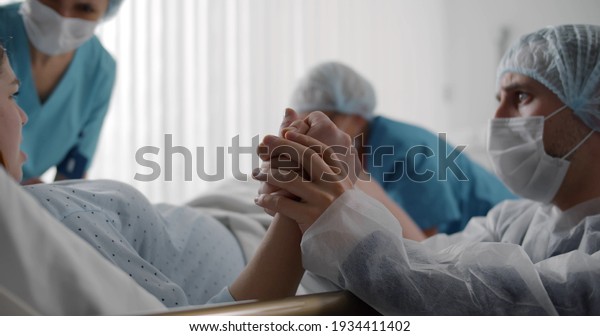 Husband in protective mask\
comforting pregnant woman during labor in ward of hospital. Young\
woman pushing giving birth in clinic delivery room with doctors and\
husband