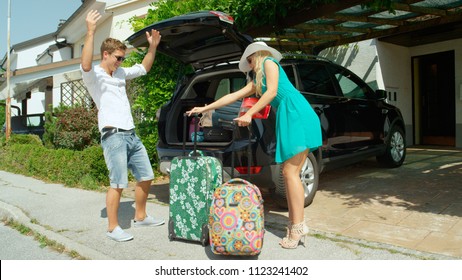 Husband Standing By The Trunk Of Their Car Can't Believe His Wife Packed Too Much Luggage For Their Road Trip. Funny View Of Young Man Frustrated With His Excited Blonde Haired Wife For Overpacking.