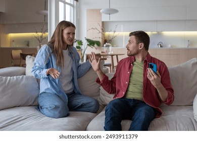 Husband and wife arguing, gesturing wildly and shouting on sofa. Wife suspects husband of infidelity, man hides phone and chats in messenger. Mistrust and tension in romantic relationships - Powered by Shutterstock