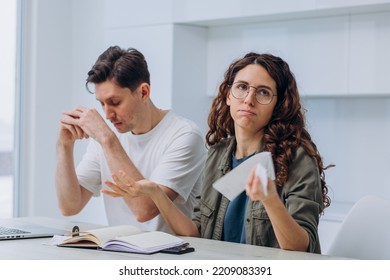 Husband and wife calculate the expense of finances sitting at home in the kitchen at the table and write it down in a notebook, a woman with checks in her hands makes excuses, a man gets upset - Shutterstock ID 2209083391