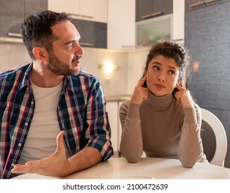 Husband and wife having argument at dining table at home. Man explaining something with dominant posture and woman holding fingers in ears and having bored expression - Shutterstock ID 2100472639