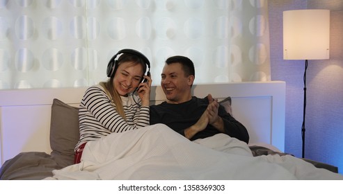 Husband And Wife In Headphones Watching TV In The Bedroom. The Family Is Having Fun And Dancing. A Man Switches Channels Using A Remote Control.