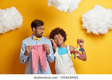 Husband and wife pose with baby items prepare for becoming parents. Cheerful pregnant woman holds mobile toy looks gladfully at man isolated over yellow background. Parenthood pregnancy concept - Powered by Shutterstock