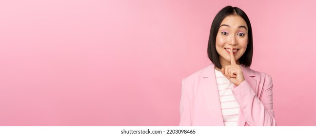 Hush, Taboo Concept. Portrait Of Asian Businesswoman Showing Shush Gesture, Shhh Sign, Press Finger To Lips, Standing Over Pink Background In Suit