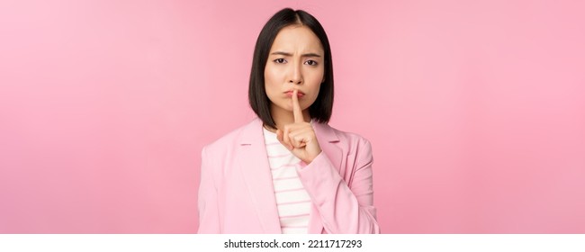 Hush, Taboo Concept. Portrait Of Asian Businesswoman Showing Shush Gesture, Shhh Sign, Press Finger To Lips, Standing Over Pink Background In Suit