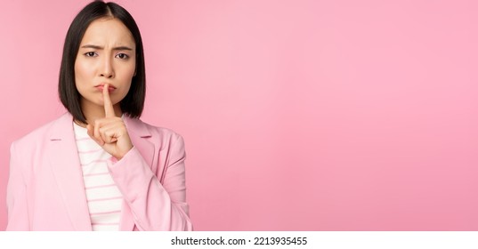 Hush, Taboo Concept. Portrait Of Asian Businesswoman Showing Shush Gesture, Shhh Sign, Press Finger To Lips, Standing Over Pink Background In Suit