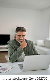 Hybrid work, elearning, online learning. Happy smiling middle aged older mature man wearing glasses looking at laptop using computer writing notes watching webinar sitting at home table, vertical.