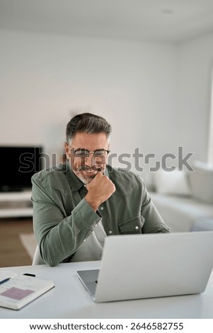 Hybrid work, elearning, online learning. Happy smiling middle aged older mature man wearing glasses looking at laptop using computer writing notes watching webinar sitting at home table, vertical.