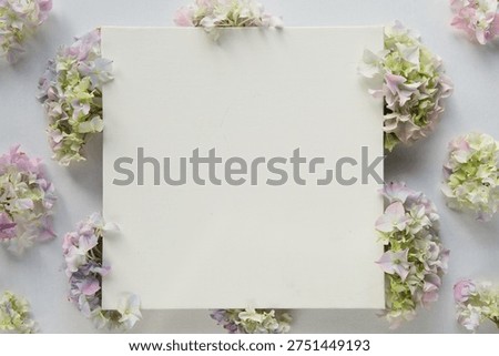 Hydrangea flowers surround blank square paper with generous central copyspace for text and design, arranged in soft pastel hues with delicate petals forming an airy flat lay composition suitable for branding and stationery