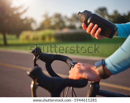 Hydrate your body. Close up of hands of professional cyclist holding water bottle, standing with his bike in park on a sunny day