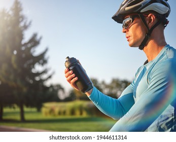 Hydration matters. Professional male cyclist holding water bottle, standing with his bike in park on a sunny day