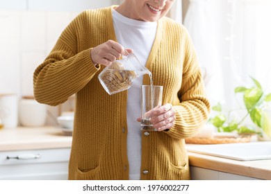 Hydration in senior ages concept. Unrecognizable grandmother standing in cozy white kitchen, pouring water into glass. Cropped of senior woman having healthy lifestyle, drinking water at home