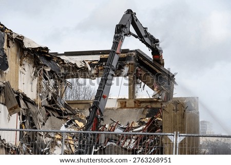 Hydraulic cracker jaws crushing and pulling down old cement office building, spraying water to control dust and debris, urban renewal demolition and construction project
