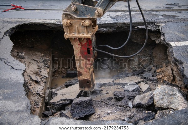 Hydraulic jackhammer from\
excavator construction truck drilling concrete ground on a busy\
asphalt road surface on which cars drive. Broken hole filled with\
muddy water.