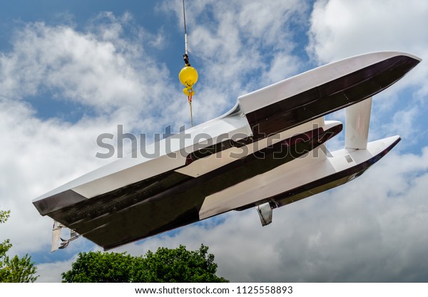 Hydroplane racing boat being\
lifted out of the water and moved overhead against a cloudy blue\
sky.