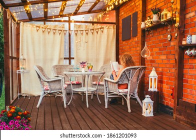 Hygge Balcony Concept. Woman Reading A Paper Book On Home Balcony Outdoors. Party String Lights Illuminating The Space. Lot Of Candles And Lanterns, Curtains Hanging With Rustic Heart Party Flags.