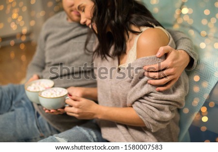 hygge, christmas and holiday concept - close up of happy couple drinking hot chocolate with marshmallow at home over shimmering festive lights