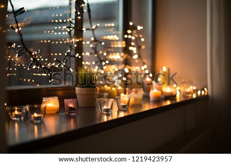 hygge, decoration and christmas concept - candles burning in lanterns on window sill and festive garland string at home