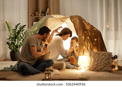 Hygge And People Concept - Father With Torch Light Telling Scary Stories To His Daughter And Wife, Family Having Fun In Kids Tent At Night At Home