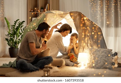 Hygge And People Concept - Father With Torch Light Telling Scary Stories To His Daughter And Wife, Family Having Fun In Kids Tent At Night At Home