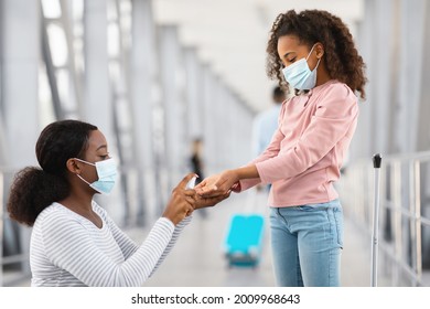 Hygiene Concept. Side View Profile Portrait Of Young Black Mum In Surgical Disposable Mask Applying Antibacterial Sanitizer Spray On Girl's Hands, Teen Cleaning Dirty Arms In Public Place At Airport