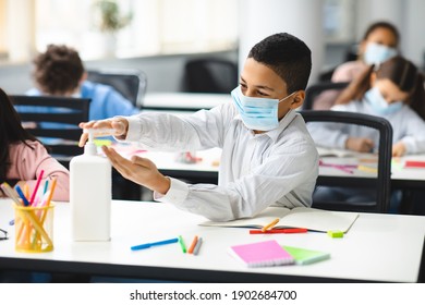 Hygiene And Health Concept. Small Schoolboy Applying Antibacterial Sanitizer On Hands From Bottle On The Desk, Wearing Protective Medical Mask, Sitting In Classroom At School. Prevent Spread Of Germs