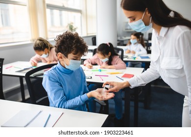 Hygiene And Healthcare Concept. Teacher Applying Antibacterial Sanitizer Spray On Small Boy's Hands, Wearing Disposable Face Mask In Classroom, Working At School. Prevent Spread Of Infection