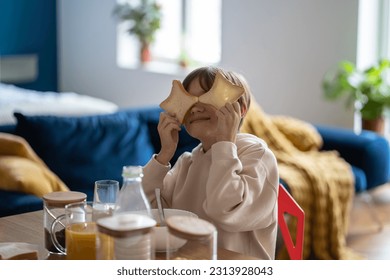 Hyperactive boy fooling around playing with pieces bread while sits at table in kitchen home. Breakfast with children. Food, good mood, cheerful playful child having fun while eating. Happy childhood