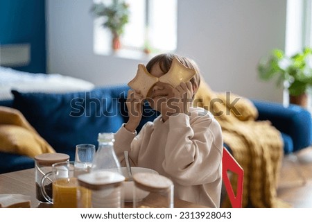 Hyperactive boy fooling around playing with pieces bread while sits at table in kitchen home. Breakfast with children. Food, good mood, cheerful playful child having fun while eating. Happy childhood