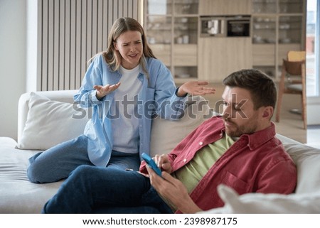 Hysterical excited woman blaming, shouting at uncaring indifferent lazy man sitting on sofa with smartphone. Wife regrets of wedding, creating family because husband doesnt satisfy expectations