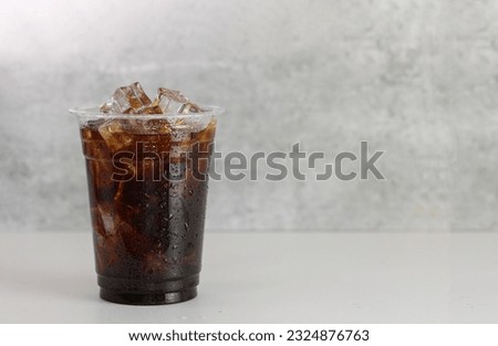 Ice americano coffee in a tall disposable glass over grey background on white table. Cold summer drink on dramatic grey background with copy space. 