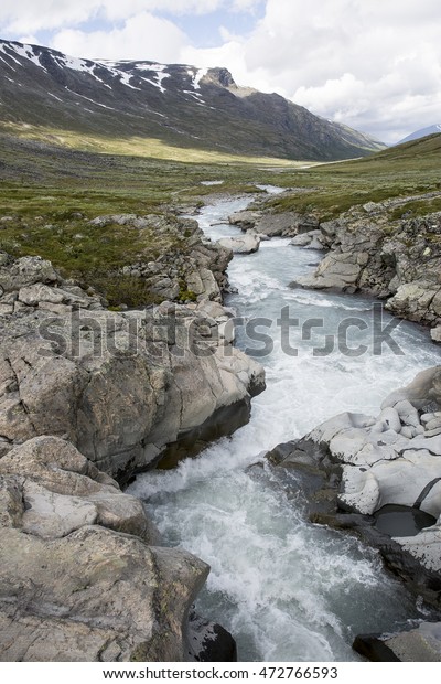 Ice blue river floating\
through a green and grey valley in the majestic mountains. Blue sky\
and white clouds in the background. The river is dividing the\
landscape in two.