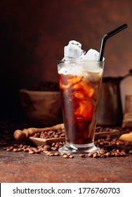 Ice Coffee With Cream Being Poured Into It Showing The Texture And Refreshing Look Of The Drink. Frozen Glass And Coffee Beans On A Old Brown Table With Kitchen Utensils. Copy Space.